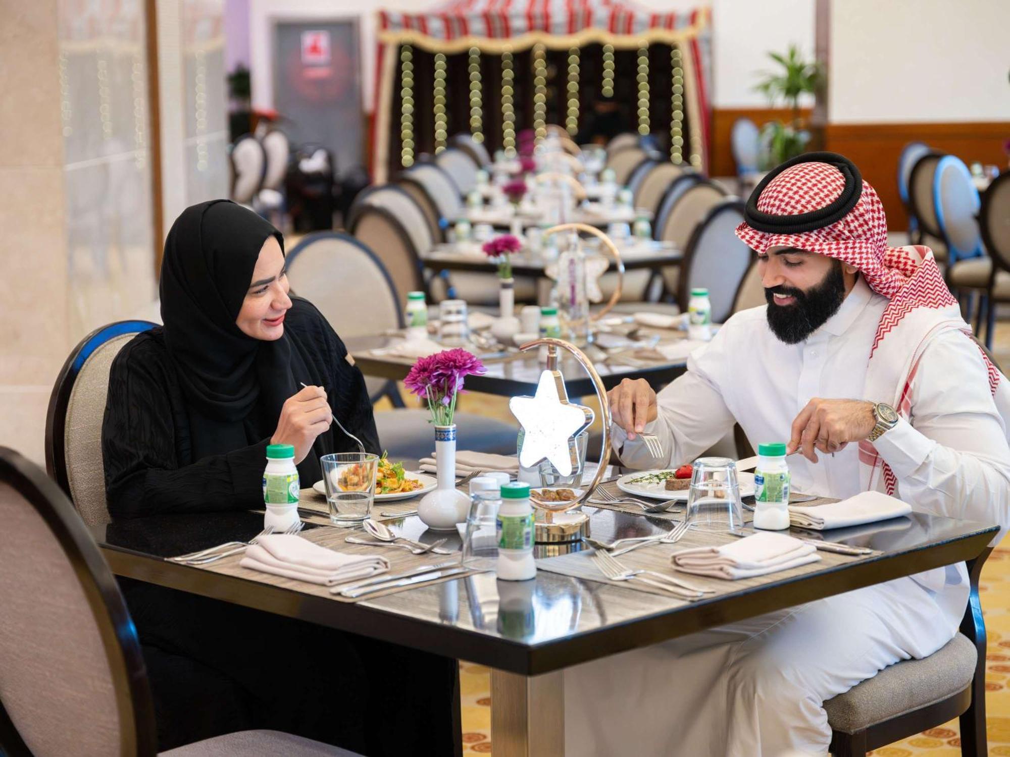 Swissotel Al Maqam Makkah La Meca Exterior foto The photo shows a couple dining at a restaurant. The man is wearing traditional attire, including a white thobe and a red and white checkered headscarf, while the woman is dressed in a black abaya and a headscarf. They are engaged in a pleasant conve