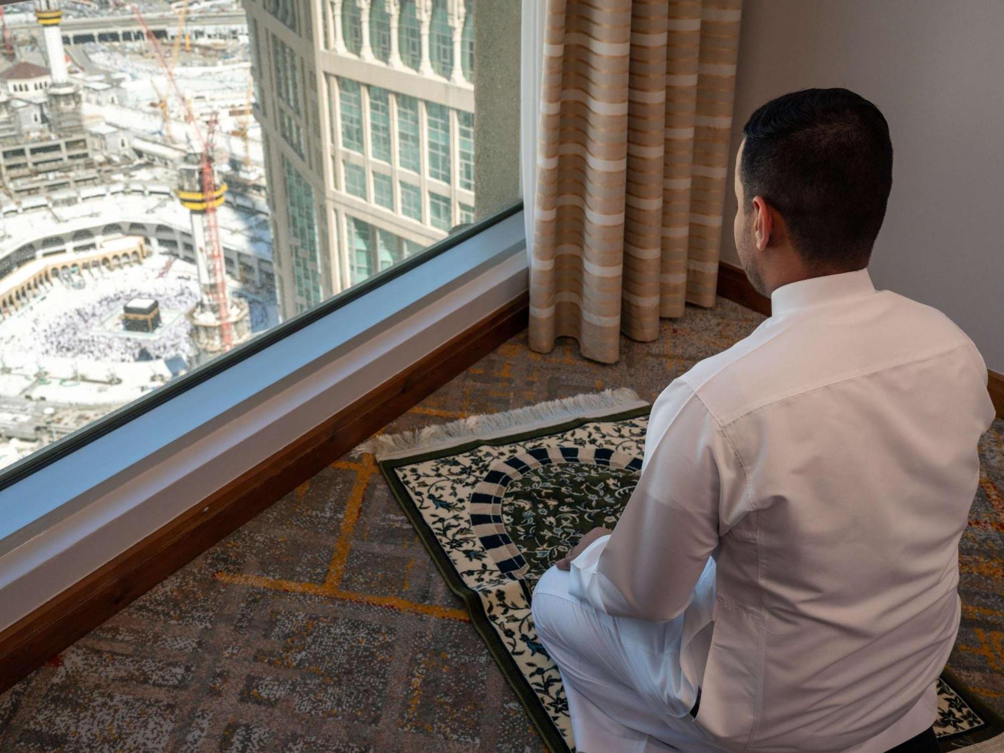 Swissotel Al Maqam Makkah La Meca Exterior foto The photo shows a man sitting in a prayer position on a prayer mat, facing a large window. Through the window, there's a view of a construction site, possibly near a significant religious site, given the context. The man is wearing traditional attire