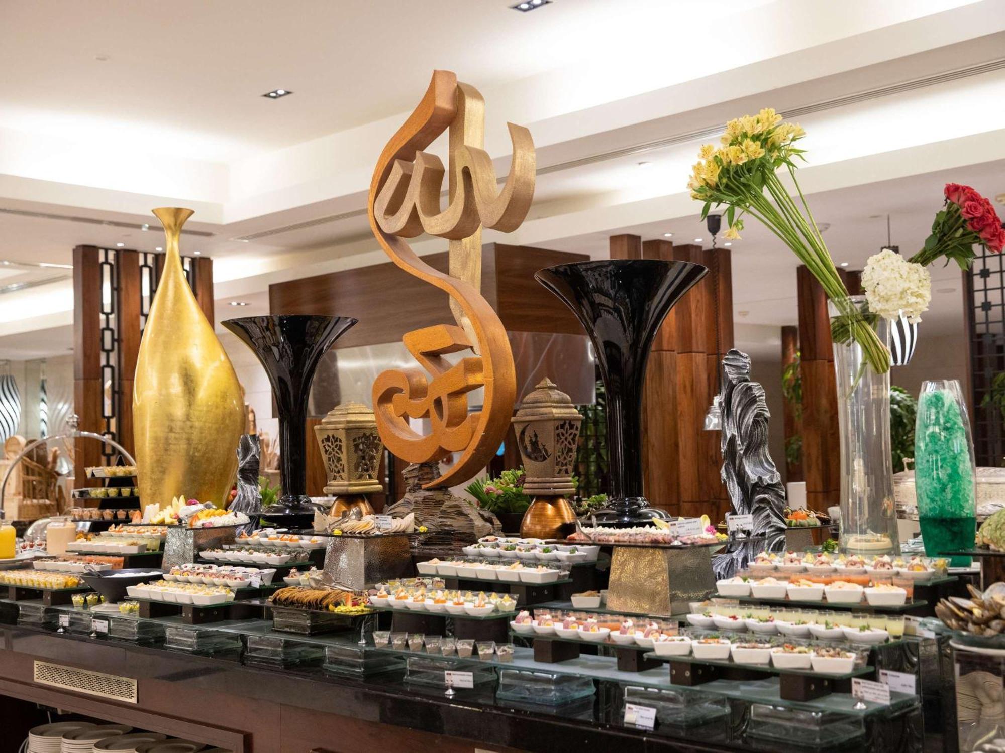 Swissotel Al Maqam Makkah La Meca Exterior foto The photo shows a beautifully arranged buffet display in a restaurant. In the center, there is a large decorative piece featuring Arabic calligraphy. Surrounding it are various elegant food presentations, including trays of desserts, likely reflectin