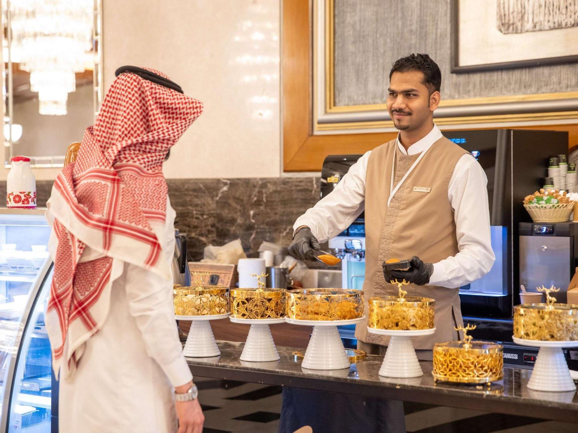 Swissotel Al Maqam Makkah La Meca Exterior foto The photo depicts a scene in a bakery or dessert shop. There is a display of various beautifully decorated cakes arranged on stands. A man wearing traditional attire, possibly a thobe and a headscarf, is interacting with a staff member who is smiling