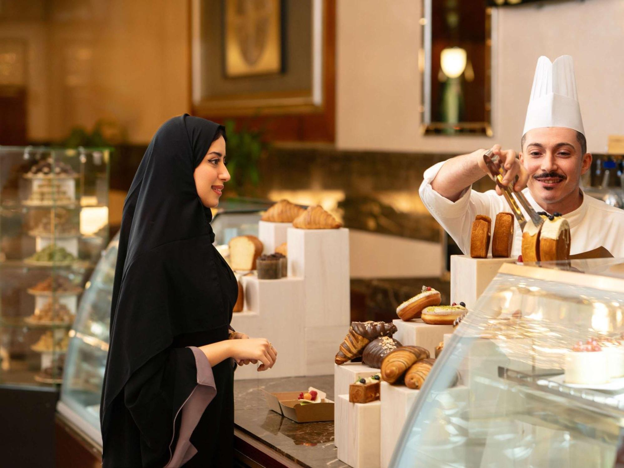 Swissotel Al Maqam Makkah La Meca Exterior foto The photo shows a bakery or pastry shop scene. On the left, a woman is wearing a black abaya and appears to be interacting with a male baker. The baker, dressed in a white chef's hat and uniform, is behind a display counter filled with various baked 